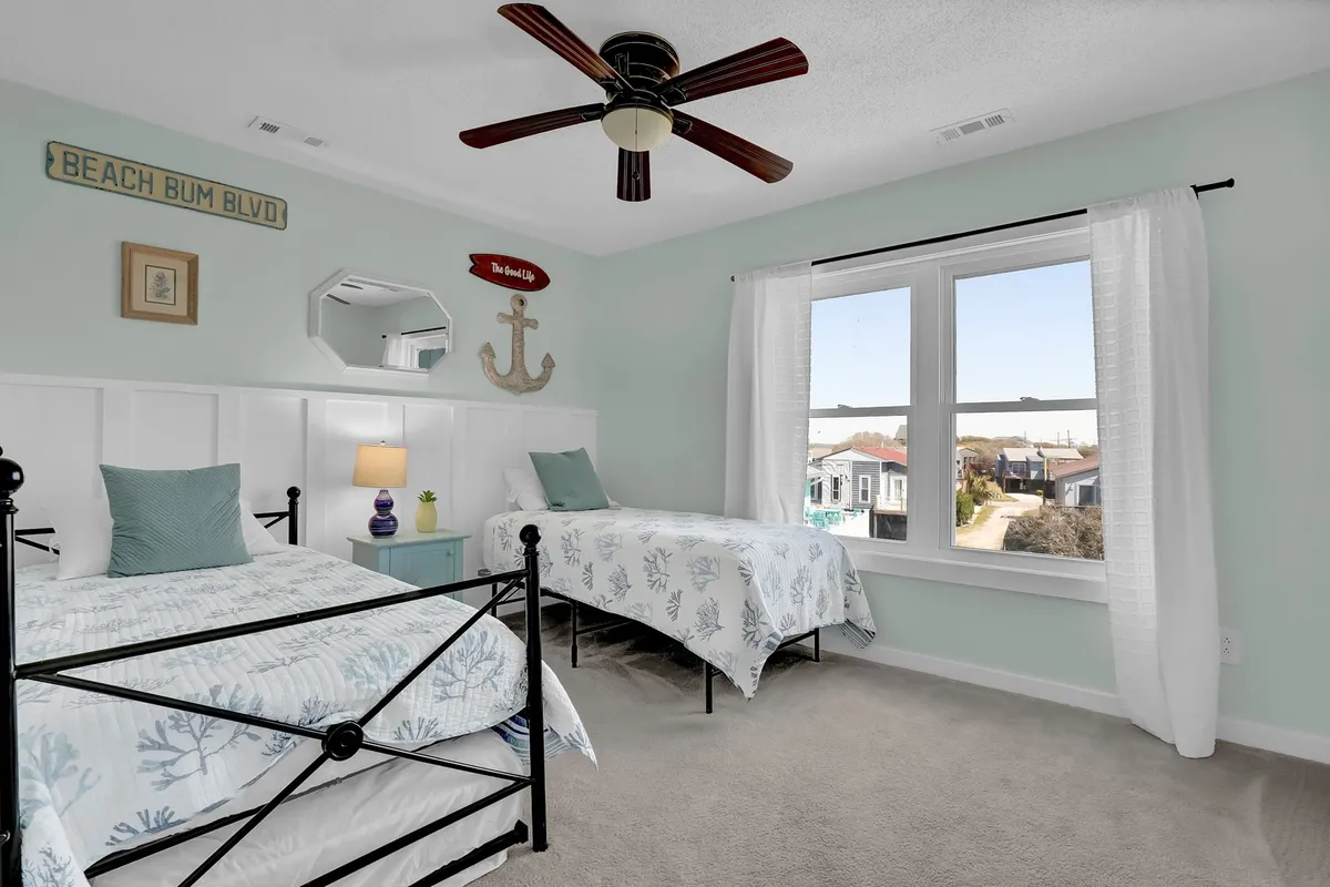 A lighthearted, inviting bedroom featuring two twin beds with beach-themed bedding, a decorative anchor, a vibrant bedside lamp, and a window offering natural light. The room feels playful and comfortable, ideal for kids or friends.