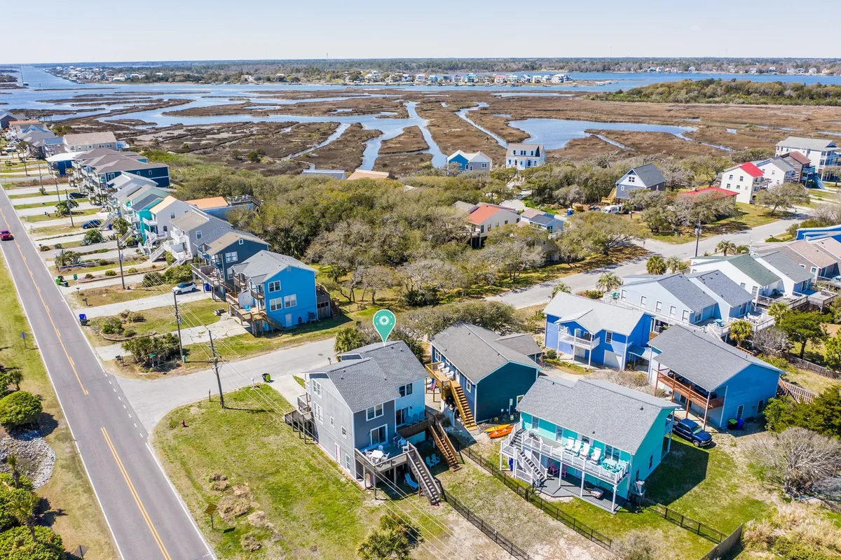 A picturesque street scene with colorful houses lining a waterfront, marshland in the distance, and ample space for strolling, biking, or exploring. The atmosphere is relaxed, with a strong sense of place and local vibrancy.