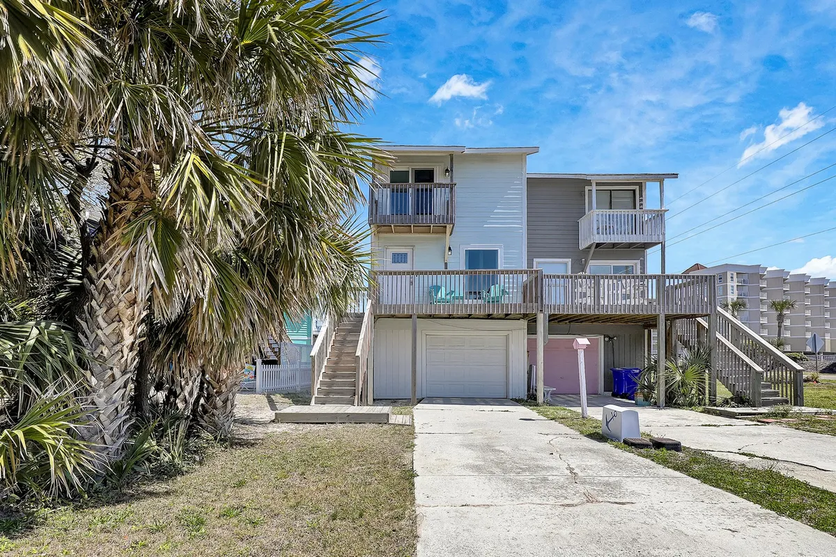 This exterior image showcases the property’s garage space, clearly visible beneath the house, with stairs leading up to the main living area. The scene includes palm trees and a welcoming balcony, giving a sense of secure parking and immediate access for unloading or transitioning from the beach.