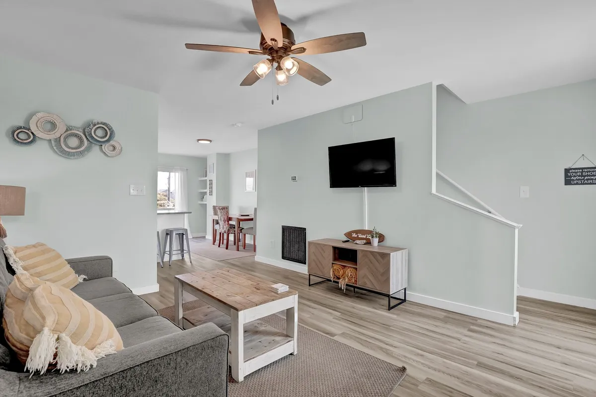 A spacious, welcoming living room with a plush sofa adorned with decorative pillows, a wooden coffee table, a ceiling fan above, and a wall-mounted TV. In the background, the dining area is visible, showcasing a seamless open layout. Large windows allow sunlight to flood the room, creating a bright and airy atmosphere.