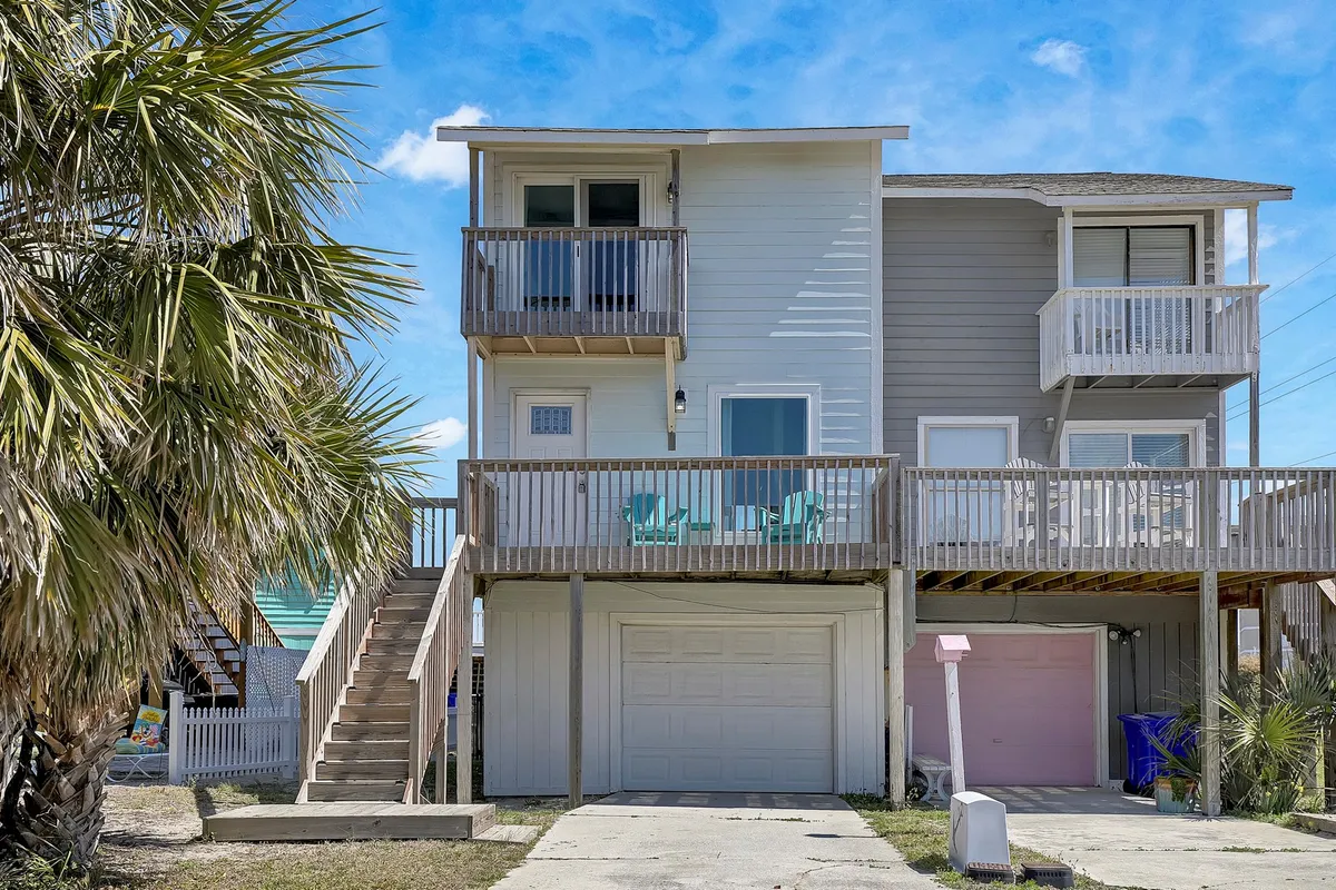 A balcony with comfortable chairs is framed by palm trees and a garage door below, suggesting a higher vantage point for relaxation. The scene communicates a sense of openness and the potential for ocean breezes, with plenty of outdoor seating for socializing or enjoying the view.