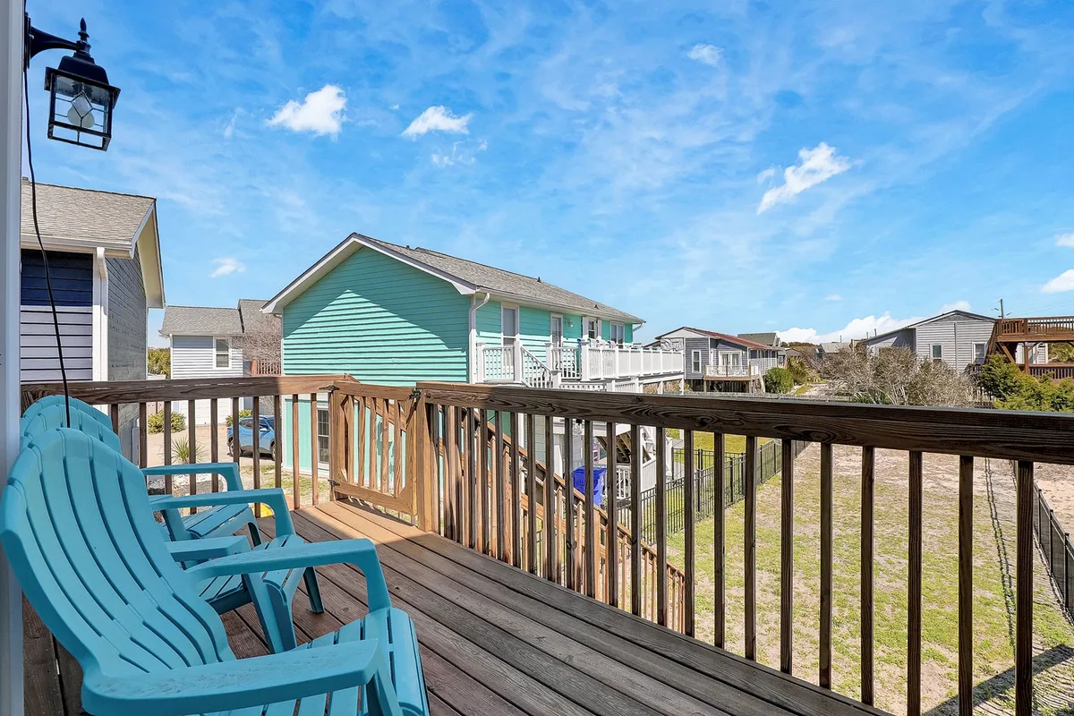 This exterior image shows a sunny wooden deck with bright blue Adirondack chairs and a green house in the background. The view suggests a private, inviting space perfect for relaxing outdoors while being able to see a portion of the yard, which could be fenced. The scene feels casual and family-friendly, evoking the idea of enjoying time outside with pets roaming safely nearby.
