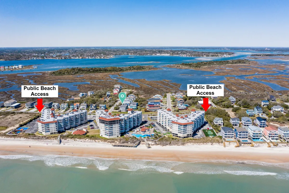 A view of public beach access signs, bordered by residential buildings and a scenic marshland stretching toward the distant ocean. Warm, late-afternoon light enhances the inviting atmosphere, while visible pathways highlight how close the home is to the beach and natural beauty.