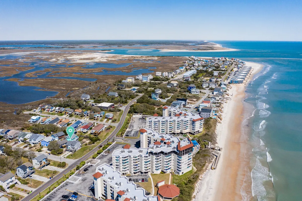 The image showcases a picturesque North Topsail Beach shoreline at sunset, with gentle waves meeting the sand, colorful coastal homes just steps from the water, and tranquil marshlands teeming with local wildlife. The relaxed atmosphere is highlighted by people strolling along the beach, enjoying the open sky and scenic vistas that extend across the ocean and marsh.